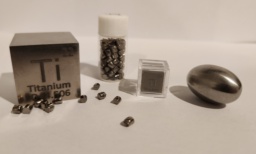 Multiple forms of Titanium metal: a 1 inch polished cube, tiny cylinders spread around and in a small bottle, a 1 cm cube in a clear case, and an egg-shaped piece