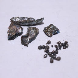 Four chunks of slightly oxidized, slightly colorful Bismuth metal, besides several tiny teardrops of Bismuth. The surface of the teardrops appear darker than the chunks.