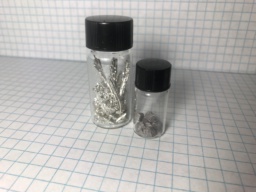 Two samples of Tin metal in its two common forms. In the bottle on the left: multiple finger-shaped crystals of shiny metallic beta-Tin, the metal that most people know about. In the small bottle on the right: small chunks of dark gray non-metallic alpha-Tin, more commonly known as Tin Pest.