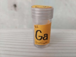 A sample of Gallium in a vial itself inside a bottle. Orange construction paper is fixed on the outer bottle with the symbol "Ga" and the electron shell.