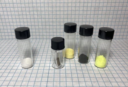 On the left: colorless |th| nitrate. On the right, in order: Tiny chunk of depleted |u| metal, Yellowcake, black Uranium dioxide, and bright yellow Uranium nitrate