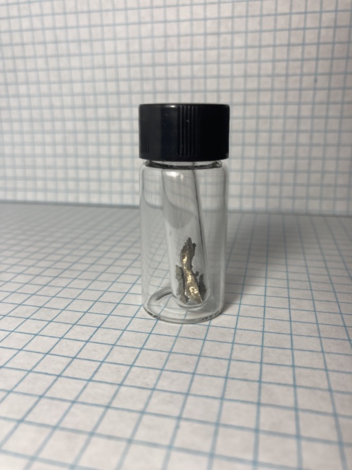 Small shiny piece of Praseodymium metal, in an ampoule which itself is in a small bottle.