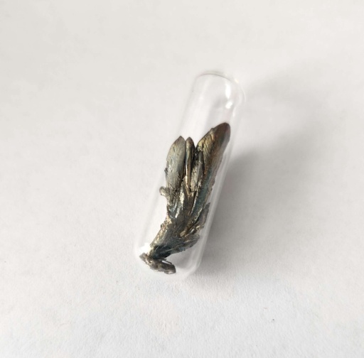 Large crystal of Barium metal sealed in an ampoule. Barium is extremely reactive and samples of this element are often dark with tarnish. This sample is shiny with a brassy sheen and a couple strips of dark oxidation. The shape of this crystal looks like a bunch of feathers, or wild onion leaves.