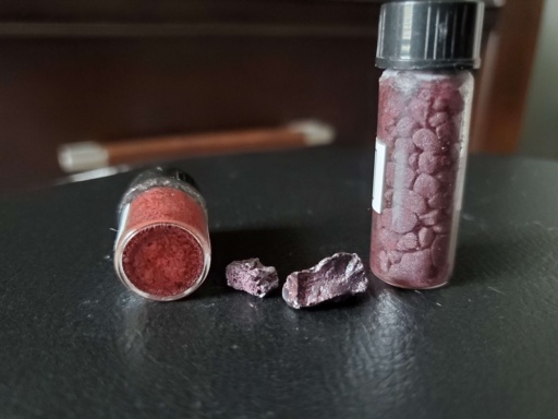 Two different allotropes of Phosphorus: a small bottle of powdery red |p|, 2 chunks of violet |p|, and a bottle filled with small chunks of violet |p|. The violet Phosphorus in this sample appears closer to a wine or light-red color.