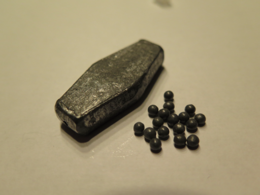 (Rephrased from contributor, and measurements are approximate.) On the left side: A small, 99% pure, mostly oxidized, coffin-shaped, 50 gram ingot of Lead. On the right side: 0.3cm diameter, 99.9% pure, completely oxidized lead shot.