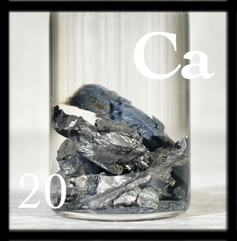 Many small chunks of Calcium metal under oil. Some surfaces are silvery, some are dull gray, and others covered in a blue layer of oxidation.